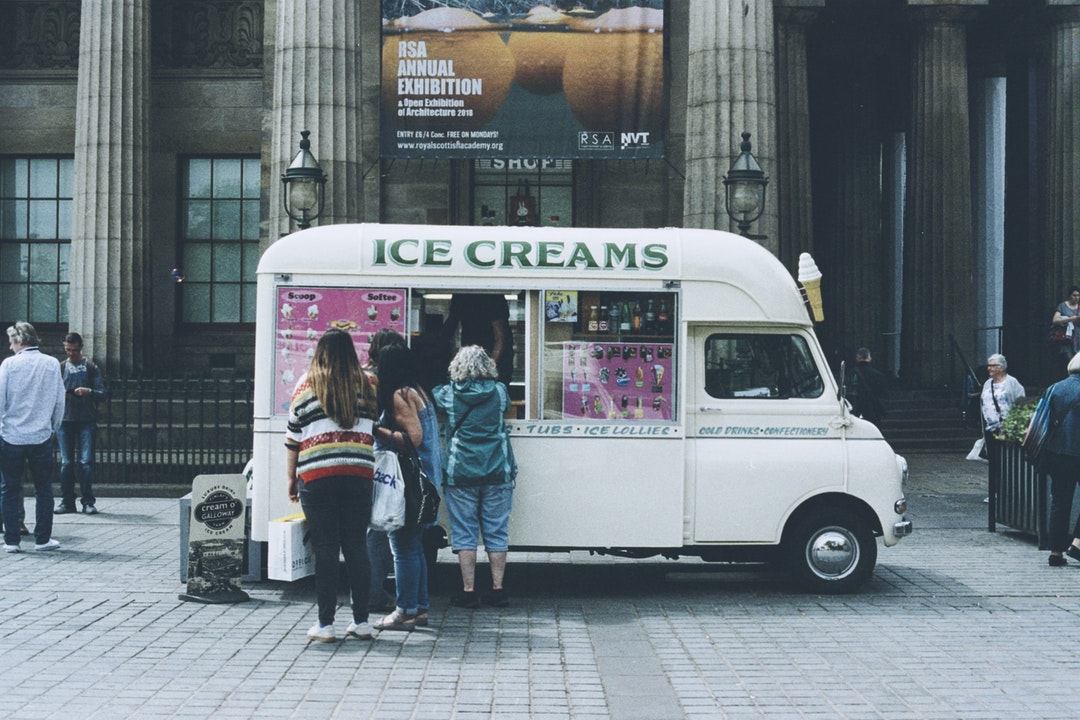 vintage looking ice cream truck with a line of customers parked in front of fancy building with pillars