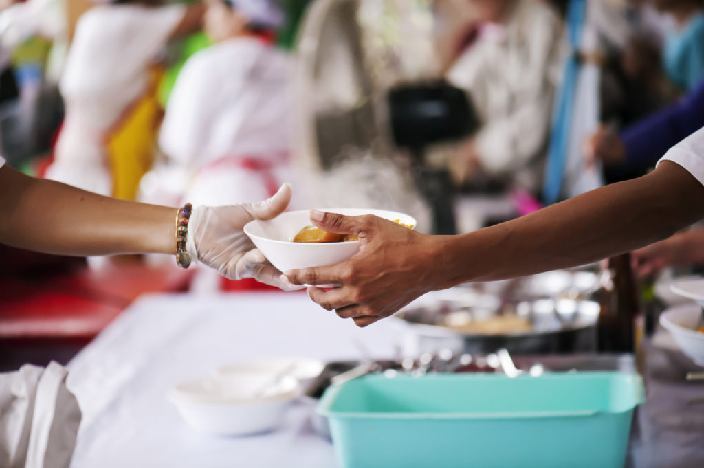 gloved hand of relief worker reaching out bowl of food to a man's hand with other people blurred in background