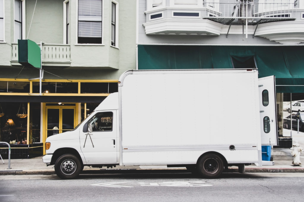 small delivery box truck parked on a city street in front of shops