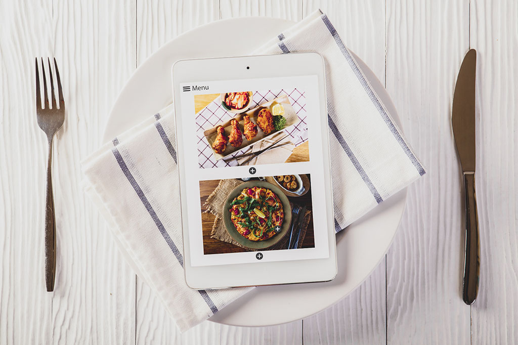 ipad with food delivery menu app on top of a white plate on a white wooden table with knife and fork on either side