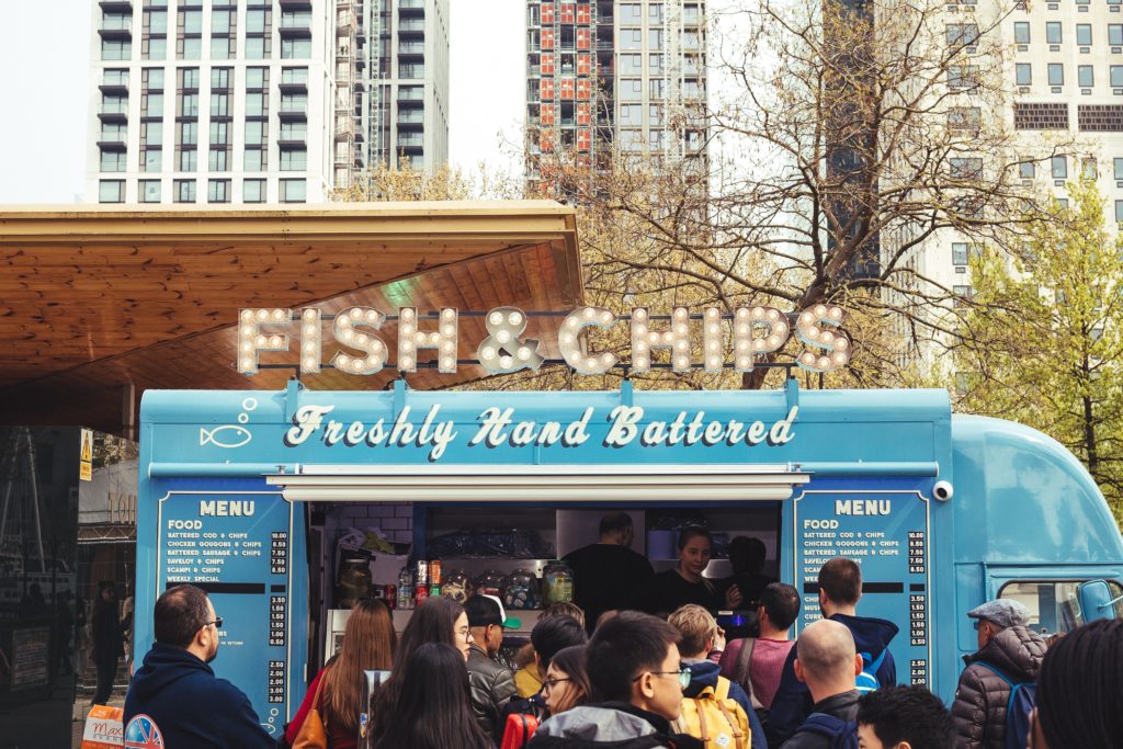 photograph of a blue food truck selling fish and chips parked in an urban area with a long line of customers