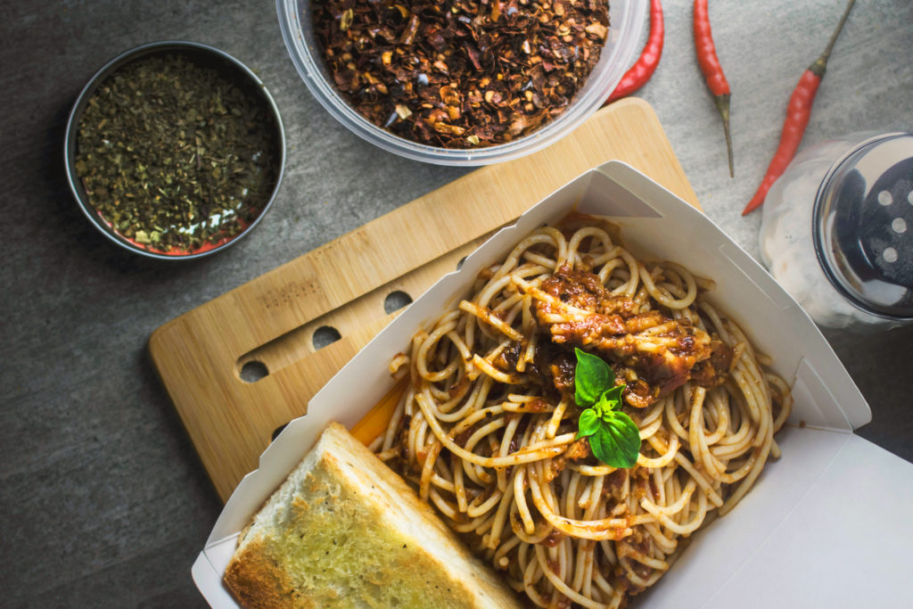 birds eye view of a tabletop with a bowl of pesto, container of red pepper spice, parmesan cheese shaker, chilis, and a food container filled with spaghetti and garlic bread