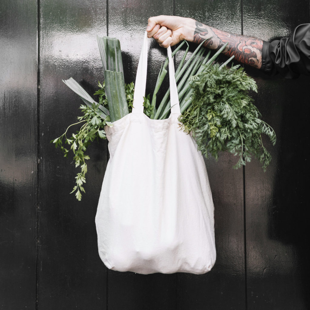 photo of a man's tattooed arm holding a tote bag full of leafy green groceries