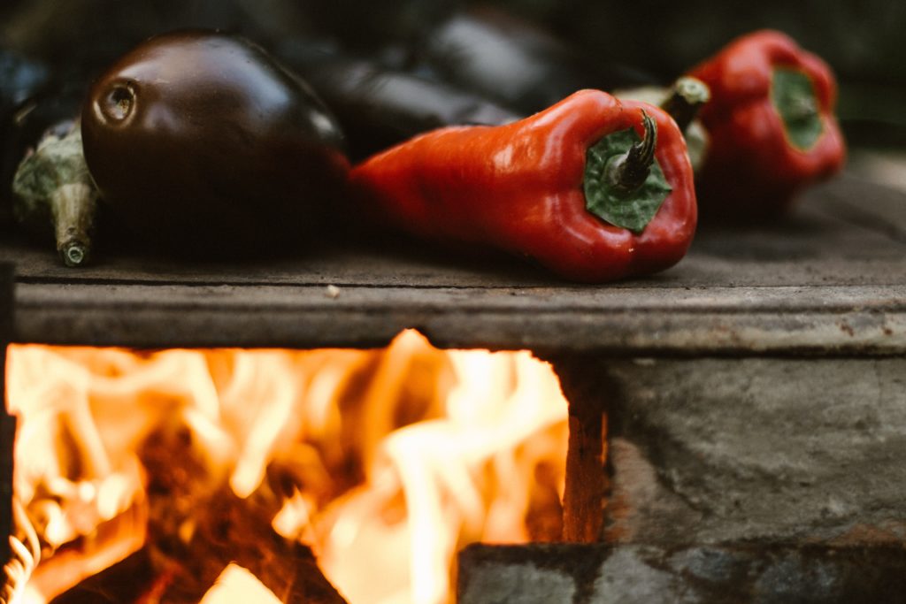 close up photo of red peppers and egg plants being roasted on a grill fire