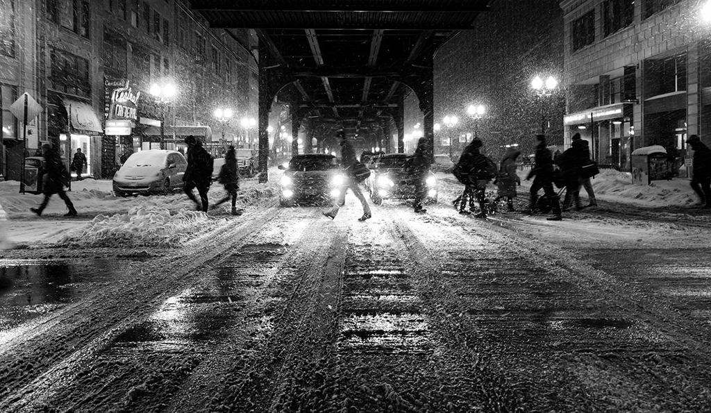 black and white photo of a snowy winter scene on urban street with pedestrians and cars